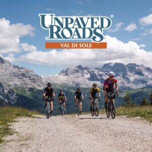 Un gruppo di ciclisti gravel pedala su una strada sterrata panoramica nelle Dolomiti, circondati da prati verdi e con maestose montagne sullo sfondo sotto un cielo azzurro con nuvole bianche. L’atmosfera è di avventura e sfida, immersi nella natura alpina.